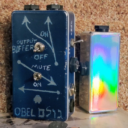 two hand-made guitar pedals. the one on the left is navy blue with white writing and arrows, three toggles and several jacks. there is a sketch of a golem character. the pedal on the right is smaller, has no controls and is covered in the same holographic rainbow vinyl as the guitar